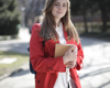 woman with a red coat, holding some books looking at the camera and wearing a backpack.