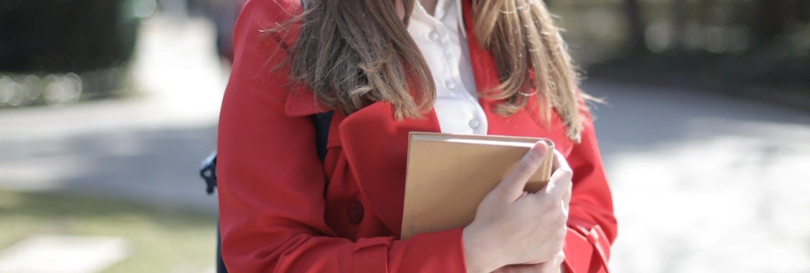 woman with a red coat, holding some books looking at the camera and wearing a backpack.