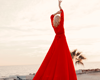 woman with her hands in the air, wearing a floor-length red dress, with her back to the camera, looking out at the ocean.