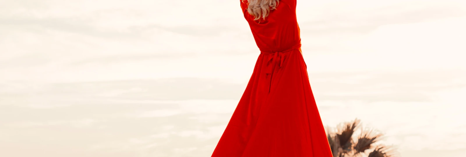 woman with her hands in the air, wearing a floor-length red dress, with her back to the camera, looking out at the ocean.