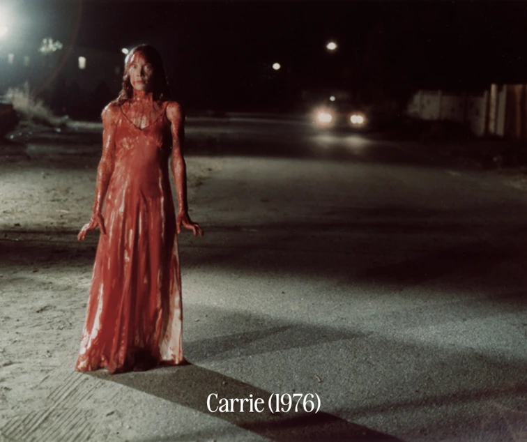 woman standing in street in long white dress, drenched head to toe in blood.