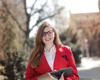 woman with a red coat, holding some books looking at the camera and wearing a backpack.