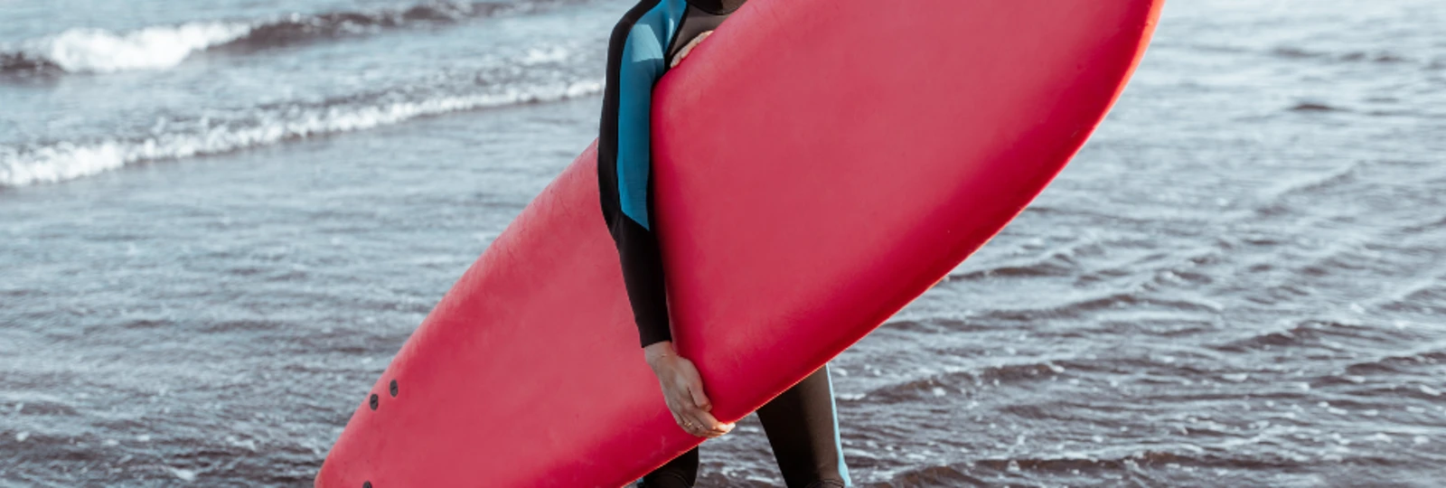 woman holding a red surfboard, wearing a wet suit and stood on the beach.