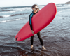 woman holding a red surfboard, wearing a wet suit and stood on the beach.