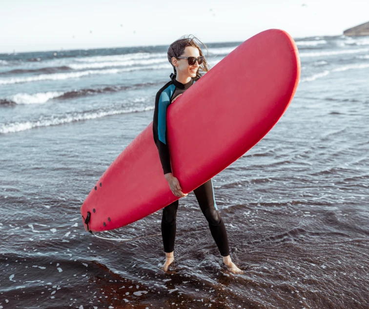 woman holding a red surfboard, wearing a wet suit and stood on the beach.