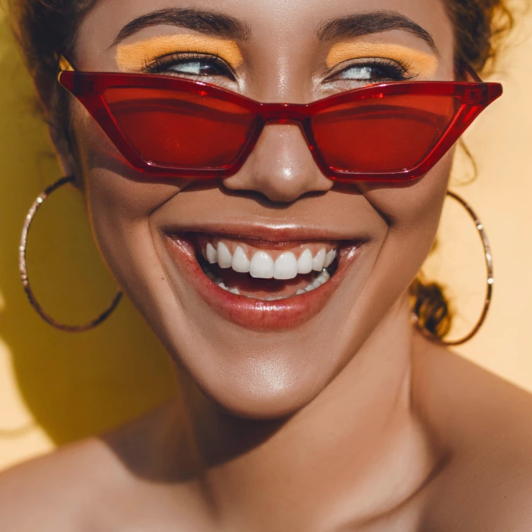 woman smiling wearing red sunglasses with bright orange eye-shadow